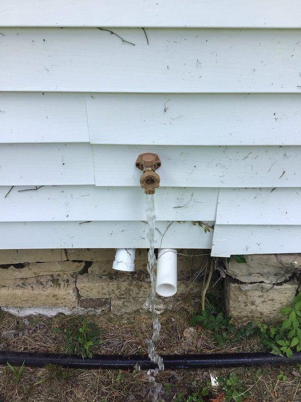 Completed free estimate for exterior water spigot replacement. Upon inspection, found existing brass hose bib actively leaking with water flowing continuously from the valve, indicating internal valve failure. Observed white PVC supply line running beneath siding to foundation level, with copper-colored fixture mounted on vinyl siding exterior wall. Documented existing conditions including protective cover on spigot, foundation deterioration with moisture staining, and black irrigation line at grade level. Photographed active water discharge and assessed plumbing system configuration for replacement recommendations.