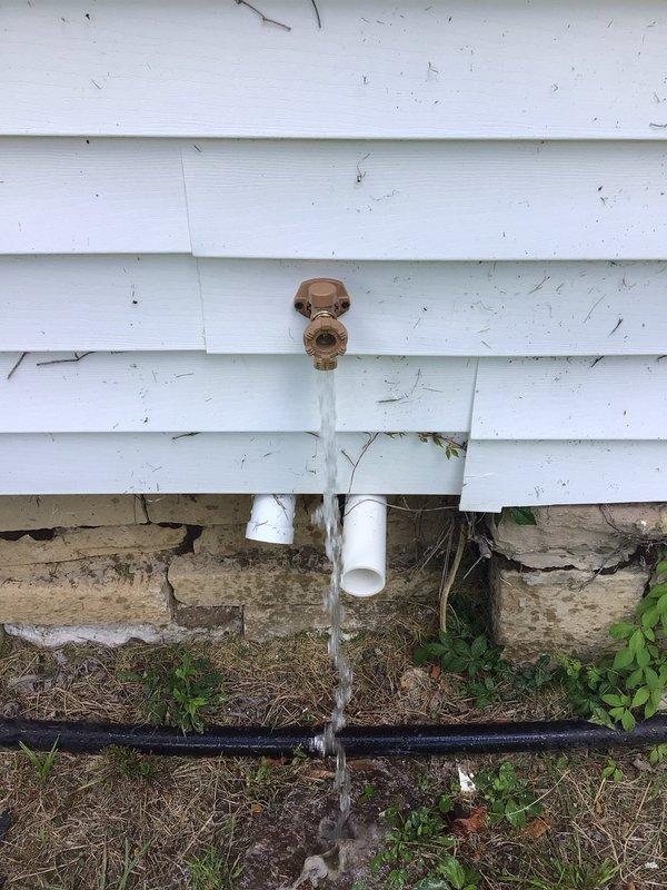 Completed free estimate for exterior water spigot replacement. Upon inspection, found existing brass hose bib actively leaking with water flowing continuously from the valve, indicating internal valve failure. Observed white PVC supply line running beneath siding to foundation level, with copper-colored fixture mounted on vinyl siding exterior wall. Documented existing conditions including protective cover on spigot, foundation deterioration with moisture staining, and black irrigation line at grade level. Photographed active water discharge and assessed plumbing system configuration for replacement recommendations.