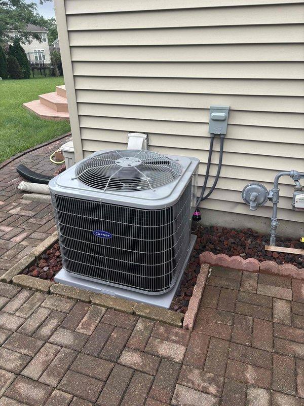Completed installation of new Carrier air conditioning system with matching evaporator coil at residential property. Outdoor condensing unit positioned on concrete pad with decorative lava rock landscaping, secured electrical disconnect box mounted to exterior wall, and refrigerant lines properly installed with black electrical conduit routed along vinyl siding. Interior work included installation of Honeywell programmable thermostat for system control. System tested and verified operational upon completion.