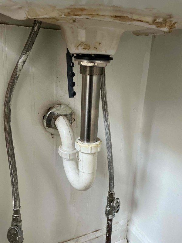 Responded to water leak complaint under bathroom vanity. Upon inspection, identified active moisture intrusion with visible water damage and rust-colored staining on cabinet interior surfaces and beadboard wall. Examined drain assembly, P-trap configuration, and supply line connections. Found compromised seal at drain body connection and deteriorated P-trap gasket allowing water seepage. Documented existing water damage patterns indicating prolonged leak condition affecting vanity cabinet and adjacent wall cavity behind laundry area. Tightened all drain connections, verified proper P-trap alignment, and inspected braided supply lines and shutoff valves for integrity. Recommended monitoring for additional leakage and potential cabinet replacement due to extent of water damage observed.