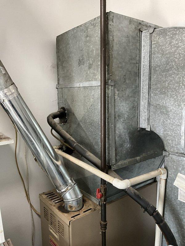 Conducted on-site assessment of existing HVAC system for replacement estimate. Documented current equipment configuration including aged air handler unit with visible surface oxidation and weathering, lower heating unit with ventilation components, and associated ductwork and piping connections. Evaluated thermostat location and existing system layout. Captured photographic documentation of equipment condition showing operational wear patterns, pipe connections, and overall system installation to provide accurate replacement specifications and sizing requirements.