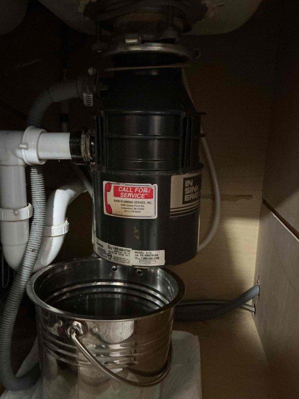 Responded to service call for non-operational and leaking garbage disposal unit that was over 20 years old. Upon inspection, found existing disposal unit showing signs of age-related deterioration with active leak present beneath sink cabinet. Placed catch bucket under unit to contain water discharge during assessment. Evaluated disposal condition, drainage connections, and mounting assembly. Due to the advanced age of the unit and combination of mechanical failure with active leaking, recommended replacement as most cost-effective solution. Documented existing installation configuration including PVC drain line connections and electrical supply for replacement specifications.