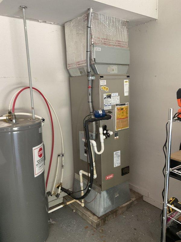 Technician responded to service call to assess existing HVAC system and provide replacement equipment pricing. Upon inspection, documented current Bryant split system consisting of outdoor cylindrical condensing unit mounted on elevated PVC platform with gravel base and indoor vertical air handler unit installed in utility space alongside water heater. System components photographed including electrical disconnect, condensate drainage piping, ductwork connections, and overall installation configuration to develop accurate replacement proposal for homeowner's review.