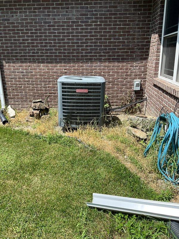 Conducted on-site evaluation of existing HVAC system experiencing humidity control issues and inadequate cooling performance. Inspected outdoor condensing unit, a Goodman-branded heat pump with cylindrical design showing significant age-related deterioration. Unit exhibited heavy organic growth, algae accumulation, and debris on exterior grille surfaces indicating extended service life and maintenance deficiencies. Documented refrigerant line connections, electrical conduit routing, and overall installation condition. Assessed system capacity requirements for residential structure with two-tone brick construction. Photographed equipment condition including weathered housing, compromised airflow surfaces, and Honeywell wireless outdoor sensor integration. Provided comprehensive replacement system estimate addressing humidity concerns and cooling capacity shortfalls.