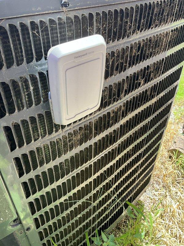 Conducted on-site evaluation of existing HVAC system experiencing humidity control issues and inadequate cooling performance. Inspected outdoor condensing unit, a Goodman-branded heat pump with cylindrical design showing significant age-related deterioration. Unit exhibited heavy organic growth, algae accumulation, and debris on exterior grille surfaces indicating extended service life and maintenance deficiencies. Documented refrigerant line connections, electrical conduit routing, and overall installation condition. Assessed system capacity requirements for residential structure with two-tone brick construction. Photographed equipment condition including weathered housing, compromised airflow surfaces, and Honeywell wireless outdoor sensor integration. Provided comprehensive replacement system estimate addressing humidity concerns and cooling capacity shortfalls.