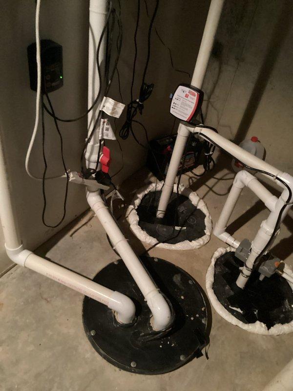 Responded to service call for sump pump system experiencing loud noises, excessive vibration, and pipe banging. Upon inspection, identified dual sump pump configuration with basement water management system including backup pump installation. Examined white PVC discharge piping network with vertical and horizontal runs, check valve assembly, and multiple connection points. Assessed electrical connections, pump basin covers, and overall system operation. Documented existing installation showing functional dual-basin setup with discharge piping routed along concrete block walls. Identified potential causes of noise and vibration issues related to pump operation and pipe movement. Provided photographic documentation of complete system configuration including piping arrangement, electrical components, and basin installations for service records.