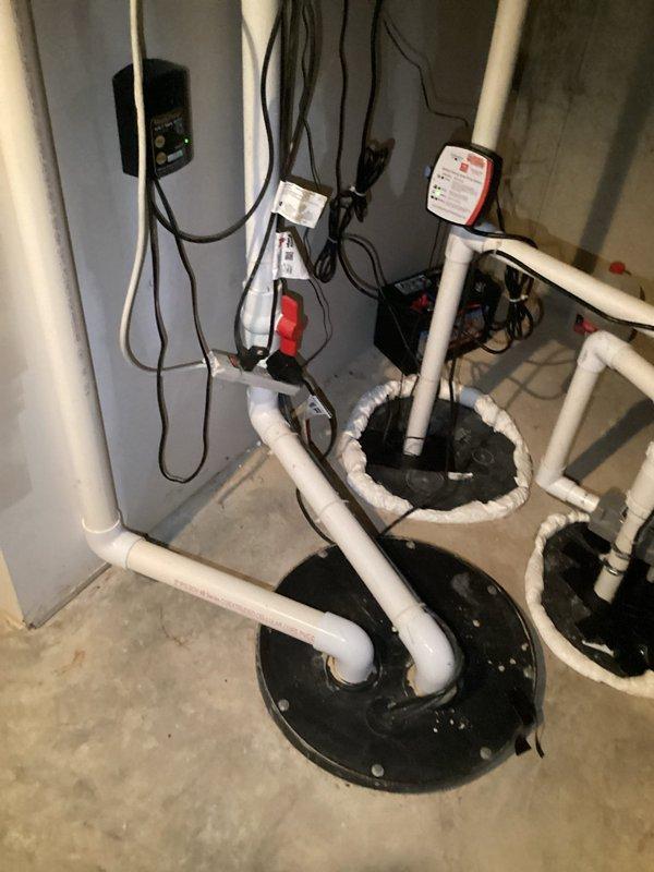 Responded to service call for sump pump system experiencing loud noises, excessive vibration, and pipe banging. Upon inspection, identified dual sump pump configuration with basement water management system including backup pump installation. Examined white PVC discharge piping network with vertical and horizontal runs, check valve assembly, and multiple connection points. Assessed electrical connections, pump basin covers, and overall system operation. Documented existing installation showing functional dual-basin setup with discharge piping routed along concrete block walls. Identified potential causes of noise and vibration issues related to pump operation and pipe movement. Provided photographic documentation of complete system configuration including piping arrangement, electrical components, and basin installations for service records.