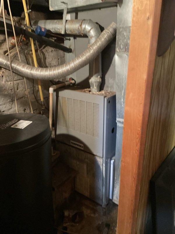 Conducted on-site evaluation of existing HVAC system for replacement estimate. Assessed aging Thompson brand gas furnace and associated mechanical components in basement installation. System currently operational but nearing end of service life. Documented existing equipment configuration including furnace unit, ductwork connections, flexible aluminum ducting, copper refrigerant lines, and plumbing infrastructure. Photographed installation layout showing gas furnace with ventilation grilles, flexible duct connections, copper piping network, and auxiliary components including black cylindrical tank. Noted exposed ductwork with visible wear, surface discoloration on metal panels, and typical residential HVAC configuration in confined utility space with concrete block walls and unfinished construction. Evaluated mechanical room constraints and existing infrastructure to determine replacement equipment specifications and installation requirements.