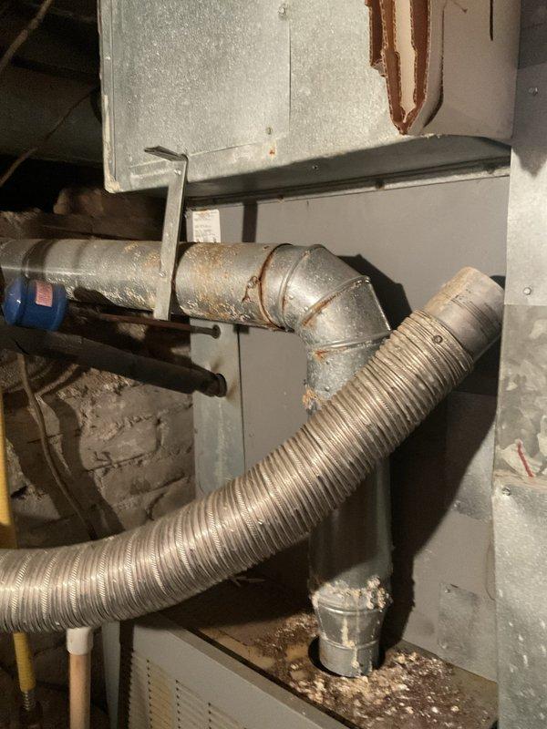 Conducted on-site evaluation of existing HVAC system for replacement estimate. Assessed aging Thompson brand gas furnace and associated mechanical components in basement installation. System currently operational but nearing end of service life. Documented existing equipment configuration including furnace unit, ductwork connections, flexible aluminum ducting, copper refrigerant lines, and plumbing infrastructure. Photographed installation layout showing gas furnace with ventilation grilles, flexible duct connections, copper piping network, and auxiliary components including black cylindrical tank. Noted exposed ductwork with visible wear, surface discoloration on metal panels, and typical residential HVAC configuration in confined utility space with concrete block walls and unfinished construction. Evaluated mechanical room constraints and existing infrastructure to determine replacement equipment specifications and installation requirements.