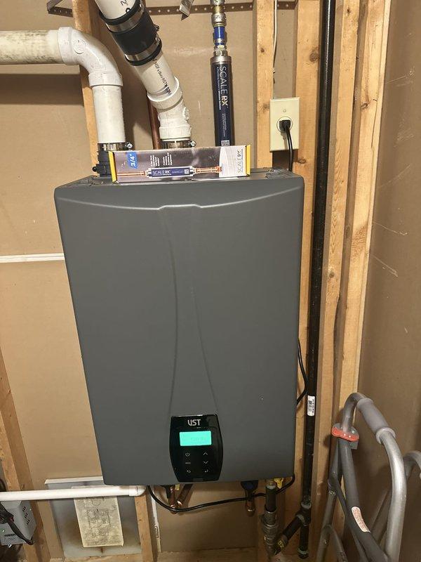 Responded to callback for tankless water heater experiencing delayed hot water delivery to second floor fixtures, with wait times of 2-3 minutes. Inspected UST tankless unit installation and verified system was configured for external recirculation capability via digital control panel. Documented existing installation showing proper venting, gas connections, copper supply lines with shut-off valves, and FlexLine connector. Confirmed recirculation pump requirement to address hot water delay issue and provided recommendations for system optimization.