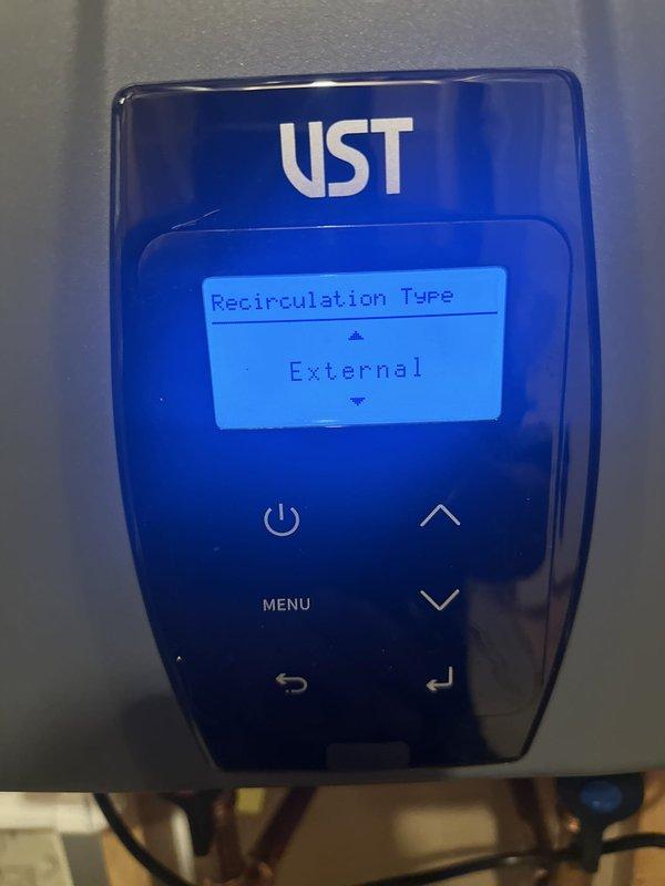 Responded to callback for tankless water heater experiencing delayed hot water delivery to second floor fixtures, with wait times of 2-3 minutes. Inspected UST tankless unit installation and verified system was configured for external recirculation capability via digital control panel. Documented existing installation showing proper venting, gas connections, copper supply lines with shut-off valves, and FlexLine connector. Confirmed recirculation pump requirement to address hot water delay issue and provided recommendations for system optimization.