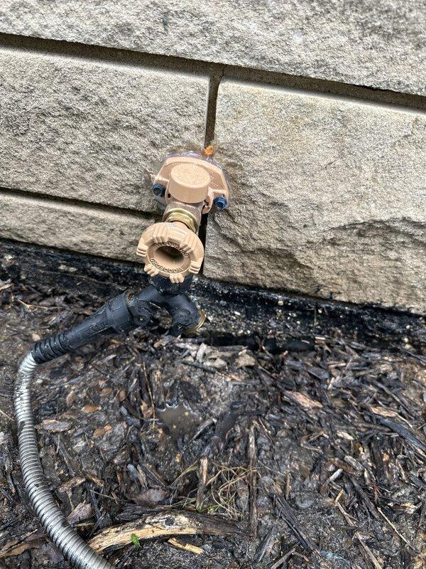 Responded to service call for outdoor hose bib with constant leak at front yard location. Upon inspection, identified failed washer and seat assembly causing continuous water discharge from spigot mounted on exterior wall with gray siding. Shut off water supply, removed existing beige plastic hose bib fixture secured with blue mounting brackets to wall. Replaced deteriorated internal components including rubber washer and valve seat. Reinstalled and secured spigot assembly to ensure proper seal against building exterior where copper supply line penetrates siding. Tested unit under full pressure to verify leak elimination and proper shutoff function. Cleared debris from ground area around installation site and confirmed no residual dripping or water discharge from repaired fixture.
