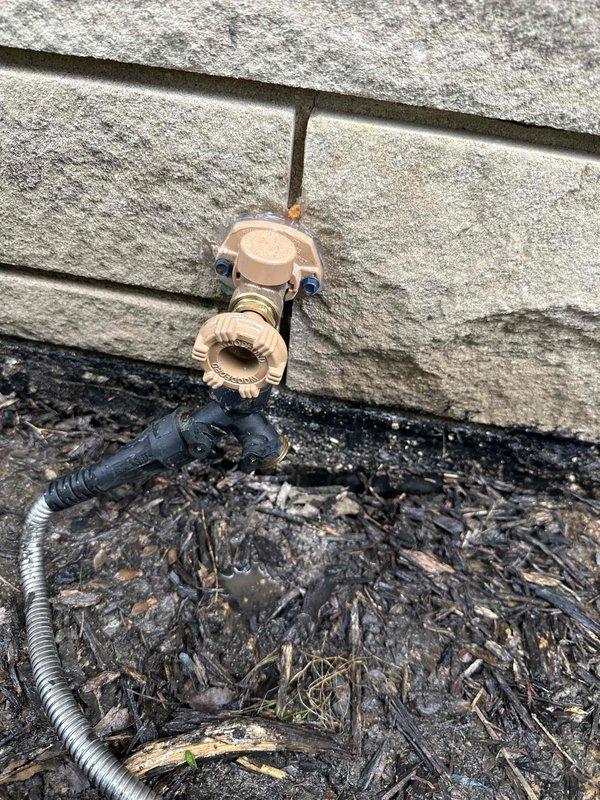 Responded to service call for outdoor hose bib with constant leak at front yard location. Upon inspection, identified failed washer and seat assembly causing continuous water discharge from spigot mounted on exterior wall with gray siding. Shut off water supply, removed existing beige plastic hose bib fixture secured with blue mounting brackets to wall. Replaced deteriorated internal components including rubber washer and valve seat. Reinstalled and secured spigot assembly to ensure proper seal against building exterior where copper supply line penetrates siding. Tested unit under full pressure to verify leak elimination and proper shutoff function. Cleared debris from ground area around installation site and confirmed no residual dripping or water discharge from repaired fixture.