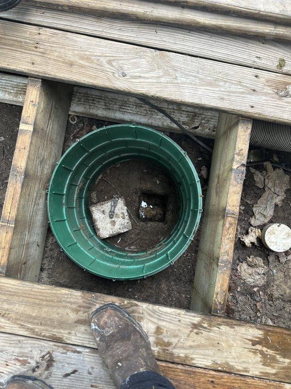 Responded to clogged drain service call and accessed underground drainage system through deck opening. Removed deck boards to expose green plastic drainage fixture and cleared blockage from beneath residential deck structure. Inspected corrugated drainage tubing for proper flow and functionality, removed debris from soil surrounding drain components, and verified system was draining properly before replacing deck access panels.