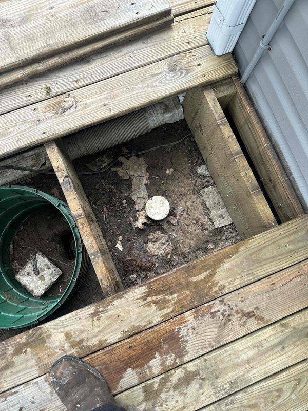 Responded to clogged drain service call and accessed underground drainage system through deck opening. Removed deck boards to expose green plastic drainage fixture and cleared blockage from beneath residential deck structure. Inspected corrugated drainage tubing for proper flow and functionality, removed debris from soil surrounding drain components, and verified system was draining properly before replacing deck access panels.