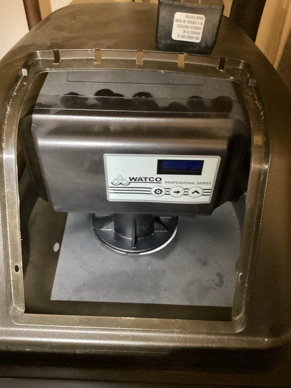 Responded to service call for non-functioning water softener with advance button malfunction and unit filled with water. Inspected water softener system and identified control valve issue preventing proper regeneration cycle. Addressed mechanical failure of advance button mechanism and assessed water level concerns within brine tank. Additionally performed requested water heater flush service, draining sediment buildup from tank bottom to improve heating efficiency and extend equipment lifespan. Documented equipment conditions with photographs showing water heater installation with drain pan, plumbing connections, and disposal unit for service records.