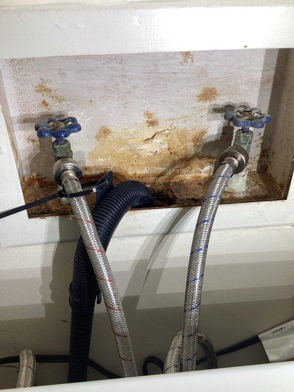 Completed annual plumbing inspection of residential property, examining all accessible water supply lines, shut-off valves, and connection points. Documented current condition of plumbing fixtures including chrome-plated flexible supply lines with blue-handled shut-off valves, noting water staining on wooden mounting panel that indicates previous moisture exposure but no active leaks present at time of inspection. All photographed components found operational and functioning within normal parameters.