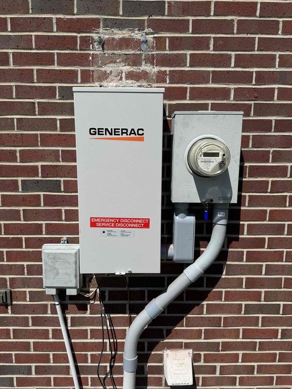 Completed full residential Generac backup generator installation including transfer switch, emergency disconnect service panel, and electrical metering system mounted to exterior brick wall. Installed white Generac transfer switch box with integrated emergency disconnect, gray utility meter housing with digital meter display, and associated junction boxes with PVC conduit runs secured to structure. All electrical connections terminated per code requirements, conduit properly supported with mounting brackets, and system tested for proper operation.