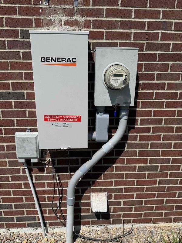 Completed full residential Generac backup generator installation including transfer switch, emergency disconnect service panel, and electrical metering system mounted to exterior brick wall. Installed white Generac transfer switch box with integrated emergency disconnect, gray utility meter housing with digital meter display, and associated junction boxes with PVC conduit runs secured to structure. All electrical connections terminated per code requirements, conduit properly supported with mounting brackets, and system tested for proper operation.