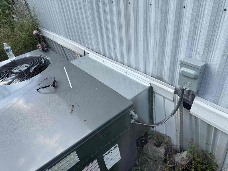 Completed installation of outdoor HVAC condensing unit positioned against corrugated metal exterior wall with proper electrical disconnect box and conduit connections. Modified return air ductwork to proper sizing per specifications and installed refrigerant line set with appropriate valve configuration. Unit was positioned in narrow service corridor with adequate clearance for maintenance access, all electrical connections secured, and system verified operational.