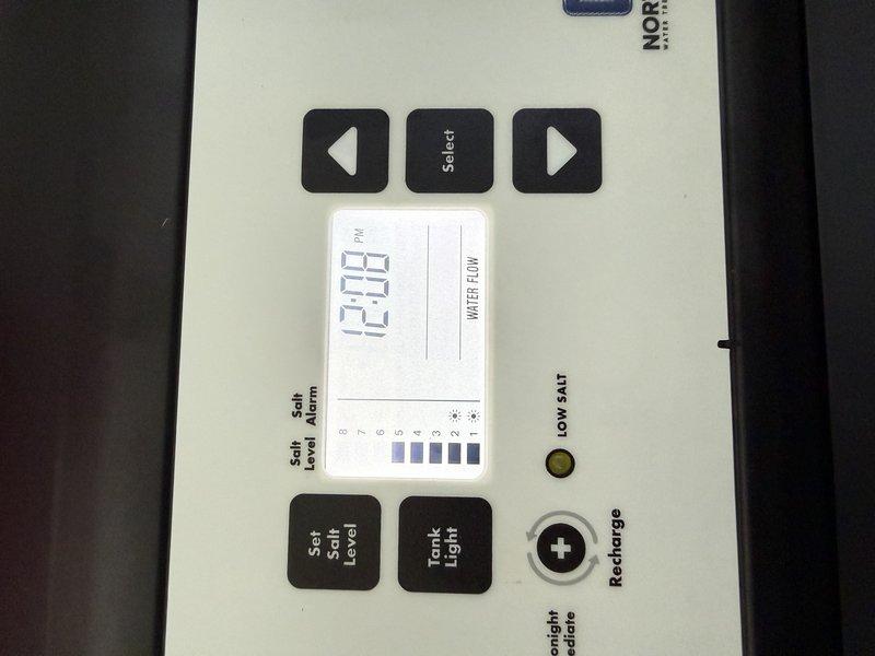 Replaced existing water softener unit with new system. Installed control panel featuring digital display with programmable settings, salt level monitoring, and manual recharge function. Connected water supply lines using braided stainless steel flex hoses with compression fittings to blue supply pipe. Mounted electronic control box beneath unit and verified all plumbing connections for proper seal and operation. Programmed system clock and tested water flow detection. Unit is operational and ready for service.