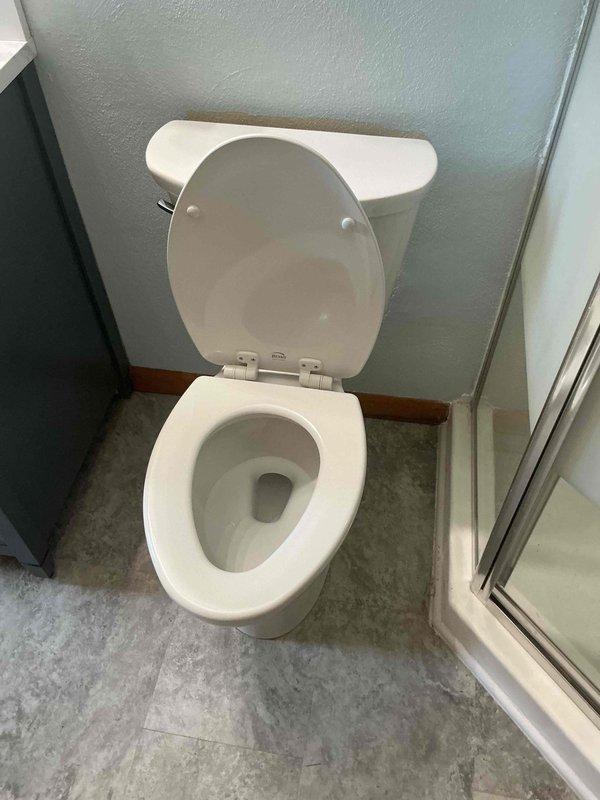 Completed toilet replacement installation in residential bathroom after previous visit identified incorrect fixture sizing. Removed existing toilet, verified proper rough-in measurements, and installed new white two-piece porcelain toilet with matching tank and bowl. Ensured proper positioning between vanity cabinet and glass shower enclosure, set fixture level on gray vinyl flooring, and secured all mounting hardware. Verified proper seal and functionality upon completion.