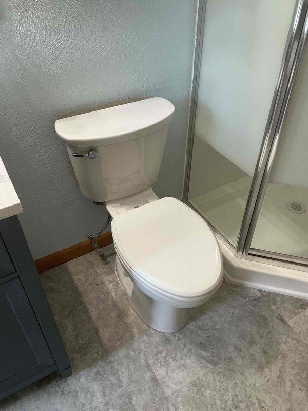 Completed toilet replacement installation in residential bathroom after previous visit identified incorrect fixture sizing. Removed existing toilet, verified proper rough-in measurements, and installed new white two-piece porcelain toilet with matching tank and bowl. Ensured proper positioning between vanity cabinet and glass shower enclosure, set fixture level on gray vinyl flooring, and secured all mounting hardware. Verified proper seal and functionality upon completion.