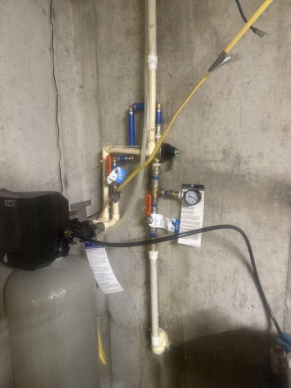 Completed site measurements for water softener reloop installation. Documented existing plumbing configuration including water treatment system with pressure gauge, brass fittings, PVC piping, and filter housing mounted on basement wall. Captured detailed photos of current setup to facilitate accurate planning and material ordering for the reloop modification.