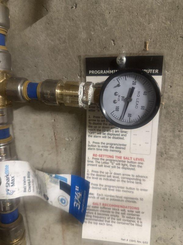 Completed site measurements for water softener reloop installation. Documented existing plumbing configuration including water treatment system with pressure gauge, brass fittings, PVC piping, and filter housing mounted on basement wall. Captured detailed photos of current setup to facilitate accurate planning and material ordering for the reloop modification.