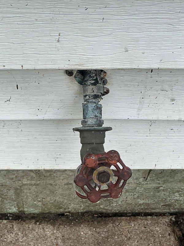 Completed outdoor spigot replacement at vacant residential property, removing heavily corroded valve assembly with damaged green fitting and rusted red wheel handle from white painted wooden exterior wall. Installed new frost-free sillcock with proper mounting through wall penetration above concrete foundation. Verified gas line connection and shut-off valve assembly at floor level near baseboard for gas stove installation, confirming proper CSST flexible gas line routing and secure connection with appropriate safety tagging in place.