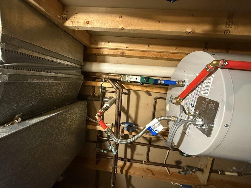 Responded to water leak complaint originating from garage area. Upon inspection, identified moisture accumulation at base of utility enclosure containing electrical components and plumbing connections. Examined water heater installation in unfinished space with exposed wooden framing and found multiple plumbing connections including hot and cold water supply lines with red and blue handles, flexible braided stainless steel connectors, and copper piping with various fittings and valves. Documented conditions with photographs showing electrical relay components, turquoise terminal blocks, and white foam insulation material around connections. Noted visible water or moisture accumulation on floor surface beneath utility enclosure. Assessed HVAC ductwork, water heater mounting, and all associated plumbing connections to determine source of leak. Investigation revealed potential leak point near water heater plumbing assembly requiring further monitoring or repair.