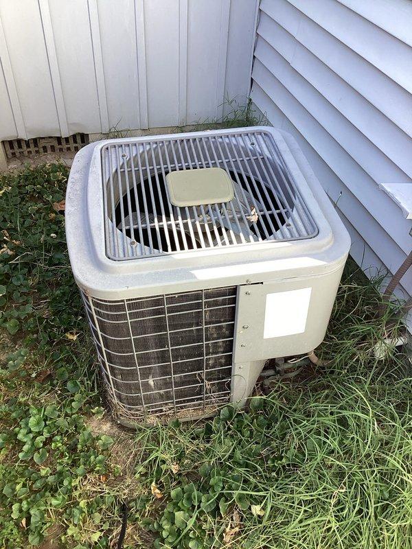 Responded to a no-cooling call with the outdoor condenser not energizing and no airflow from supply vents, and performed a diagnostic evaluation of the A/C system. Inspected the 14x20x1 return air filter and grille, noting accumulated dust and debris, and examined the condenser, which showed a dirty, debris-laden coil and fan guard with the unit sitting directly on soil without a pad, and surrounding vegetation encroaching on the required clearance.