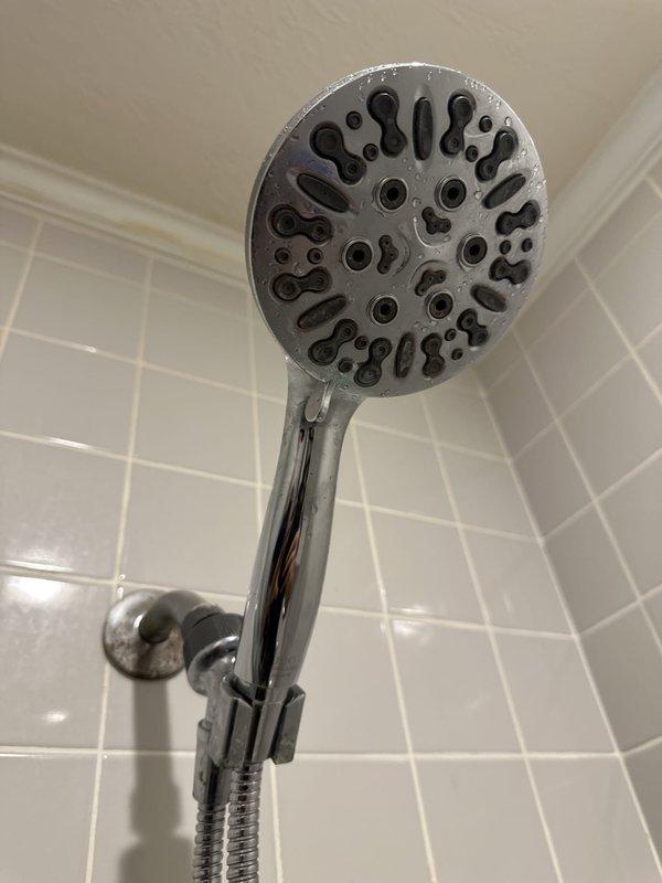 Inspected the shower for a dripping fixture and evaluated the valve assembly, accessing it from the valve trim inside the shower since no rear access panel is present. Identified the handheld showerhead, shower arm, and wall flange with visible mineral buildup and surface spotting, and noted staining on the upper wall and ceiling edge for monitoring of possible moisture intrusion. Diagnosed the hot-side leak and valve condition for repair or replacement of the shower valve components.