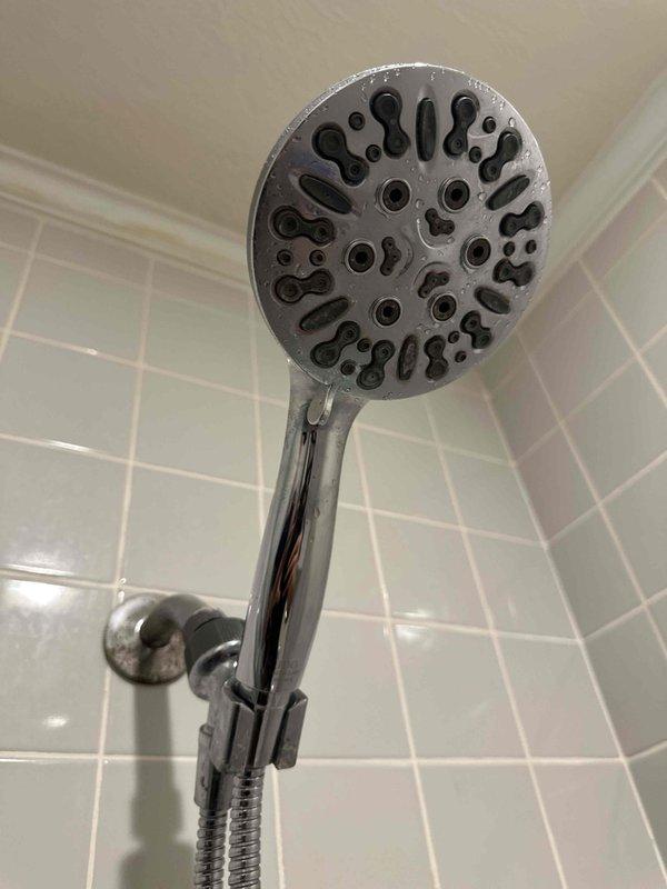 Inspected the shower for a dripping fixture and evaluated the valve assembly, accessing it from the valve trim inside the shower since no rear access panel is present. Identified the handheld showerhead, shower arm, and wall flange with visible mineral buildup and surface spotting, and noted staining on the upper wall and ceiling edge for monitoring of possible moisture intrusion. Diagnosed the hot-side leak and valve condition for repair or replacement of the shower valve components.
