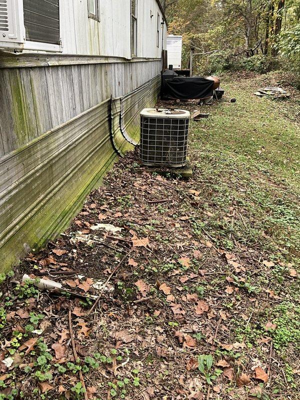 Performed an on-site evaluation for replacing an aging furnace with a heat pump system at a manufactured home, including assessment of the existing outdoor condenser, electrical disconnect, and exterior equipment placement. Documented the existing condenser, which showed dirty coil surfaces with mineral buildup, along with surrounding site conditions, to determine installation requirements and equipment sizing for the heat pump conversion.