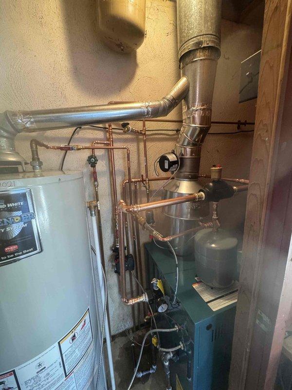 Installed a new boiler with an expansion tank mounted on top, connected via copper piping with a Y-strainer and brass fittings. Piped the system with soldered copper supply and return lines, a circulator pump, air separator, isolation valves, and a pressure gauge, and connected braided flexible lines and electrical wiring. Installed a stainless steel vent assembly and tied it into the existing flue stack alongside the gas water heater, with foil-taped joints sealed and warning labels in place.