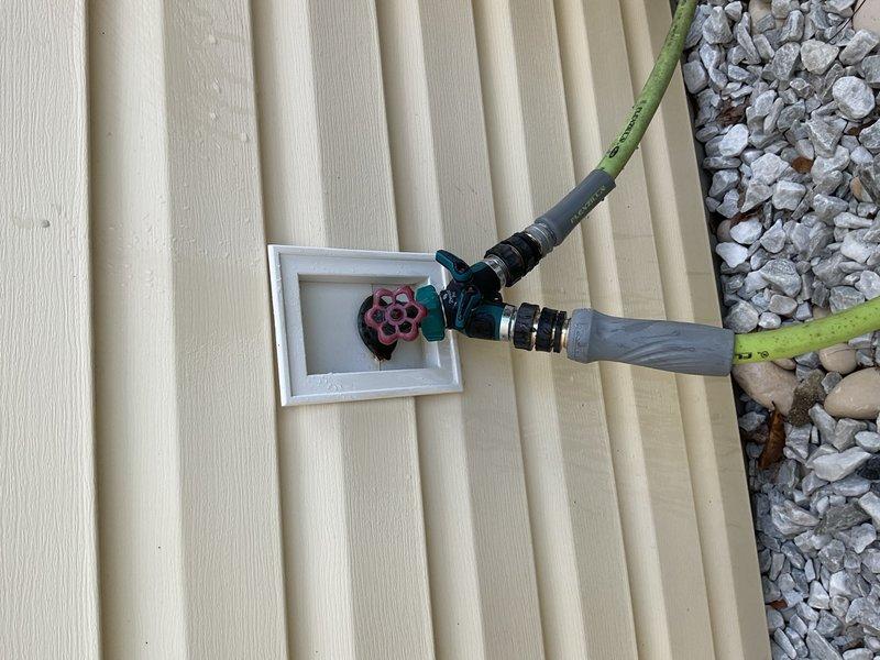 Installed outdoor water spigot system with Y-connector and dual hose setup on exterior wall with vinyl siding. Completed PVC drainage line installation at foundation level with proper gravel bed and landscape fabric for erosion control and weed prevention.
