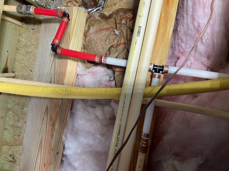 Performed inspection and documentation of residential plumbing system installation in basement crawl space area. Examined multiple utility lines including PEX water supply lines, PVC drainage piping, and gas supply connections running through floor joists. Verified proper routing of red PEX plumbing with metallic fittings, white PVC drainage system marked with specification codes, and beige gas line with T-joint configuration. Assessed condition of exposed infrastructure including wooden joists, OSB sheathing, and pink fiberglass insulation between structural members. Documented existing system layout showing parallel pipe runs through framed cavities for maintenance records and future reference.