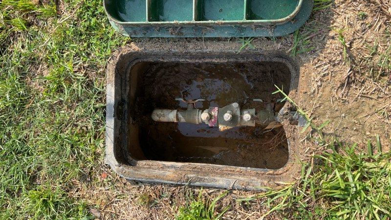 Performed photographic documentation of underground utility access infrastructure. Inspection revealed an open utility access box with standing water and submerged brass valve assembly requiring attention. Additionally documented exterior walkway surfaces including concrete pavers, artificial turf border, and natural grass areas with pressure washing equipment visible in frame for maintenance assessment purposes.