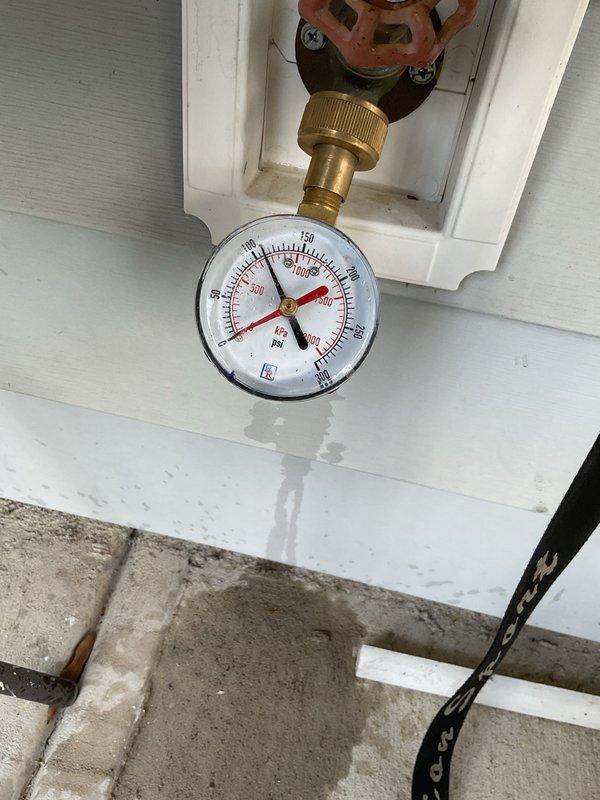 Inspected and documented exterior water supply system including pressure gauge assembly and shutoff valve mounted on building exterior. Pressure gauge reading verified and photographed showing PSI measurements and brass fittings in operational condition. Red wheel-style shutoff valve confirmed accessible and functional. 

Assessed irrigation system components in landscaped garden bed area with mulched planting zones. Located and documented circular irrigation valve cover and associated green valve component within mulch bed adjacent to concrete patio surface. Vegetation and mulch coverage observed around irrigation access points to ensure proper system accessibility for future maintenance requirements.
