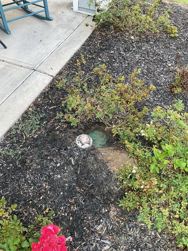 Inspected and documented exterior water supply system including pressure gauge assembly and shutoff valve mounted on building exterior. Pressure gauge reading verified and photographed showing PSI measurements and brass fittings in operational condition. Red wheel-style shutoff valve confirmed accessible and functional. 

Assessed irrigation system components in landscaped garden bed area with mulched planting zones. Located and documented circular irrigation valve cover and associated green valve component within mulch bed adjacent to concrete patio surface. Vegetation and mulch coverage observed around irrigation access points to ensure proper system accessibility for future maintenance requirements.