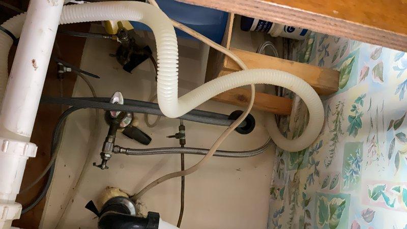 Responded to under-sink plumbing inspection and documentation. Upon arrival, accessed cabinet beneath kitchen sink and photographed existing plumbing configuration including garbage disposal unit, drainage assembly with white corrugated flexible drain line in S-curve configuration, and associated connection points. Documented standard residential plumbing fixtures, metal and plastic fittings, and flexible supply lines. Also photographed adjacent utility area showing water supply system components including mounted pump unit, flexible tubing connections, white drainage pipes, and corrugated ventilation hose. All components appeared functional at time of inspection with standard mounting and connection methods in place.