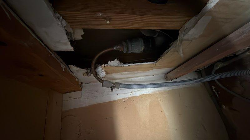 Inspected and documented plumbing infrastructure in confined crawl space beneath residential structure. Verified proper installation and operational status of drainage system components, including metal trap assembly and flexible ductwork secured to wooden framing members. Assessed condition of exposed plumbing fixtures and confirmed all connections were intact and functioning within normal parameters.