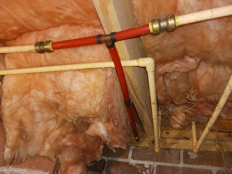 # Plumbing Installation and Crawl Space Infrastructure Work

Completed installation and routing of PEX plumbing lines in unfinished attic space with exposed framing. Installed cream-colored PEX piping with red PEX junction section, secured with brass push-fit compression fittings along wooden structural members. Routed plumbing configuration through framed cavity above brick masonry structure, working around existing pink fiberglass insulation between ceiling joists.

Performed work in crawl space beneath building, addressing utility infrastructure alongside existing systems including white PVC drain lines, black HVAC ductwork, and electrical conduits. Secured flexible tubing to floor joists using white mounting clips. Removed and cleared construction debris and displaced insulation materials from crawl space floor near foundation wall with ventilation grate. All plumbing connections properly fitted and secured to framing for permanent installation prior to final wall covering installa