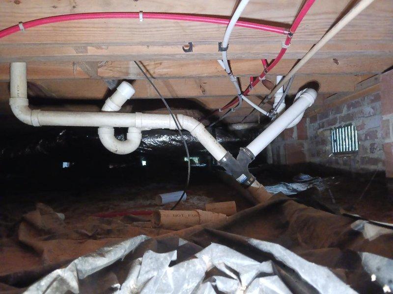 # Plumbing Installation and Crawl Space Infrastructure Work

Completed installation and routing of PEX plumbing lines in unfinished attic space with exposed framing. Installed cream-colored PEX piping with red PEX junction section, secured with brass push-fit compression fittings along wooden structural members. Routed plumbing configuration through framed cavity above brick masonry structure, working around existing pink fiberglass insulation between ceiling joists.

Performed work in crawl space beneath building, addressing utility infrastructure alongside existing systems including white PVC drain lines, black HVAC ductwork, and electrical conduits. Secured flexible tubing to floor joists using white mounting clips. Removed and cleared construction debris and displaced insulation materials from crawl space floor near foundation wall with ventilation grate. All plumbing connections properly fitted and secured to framing for permanent installation prior to final wall covering installa