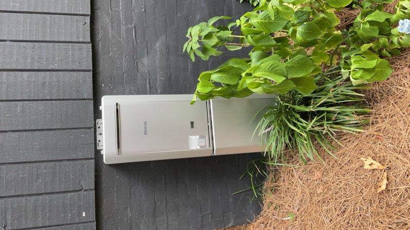 Completed installation of outdoor tankless water heater unit on exterior wall with professional mounting hardware and proper clearance from adjacent landscaping. Verified HVAC condenser unit positioning on concrete pad and confirmed adequate clearance and accessibility for both heating and cooling systems in residential side yard utility area.