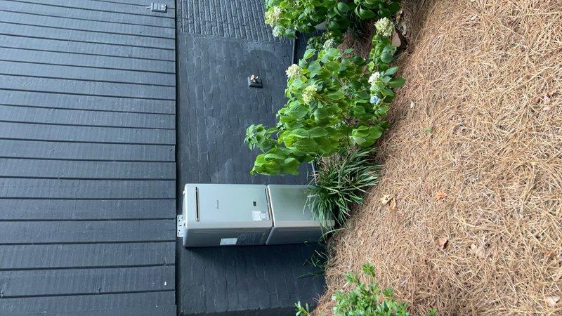 Completed installation of outdoor tankless water heater unit on exterior wall with professional mounting hardware and proper clearance from adjacent landscaping. Verified HVAC condenser unit positioning on concrete pad and confirmed adequate clearance and accessibility for both heating and cooling systems in residential side yard utility area.