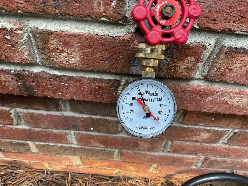Performed inspection and documentation of backflow prevention assembly and associated pressure monitoring equipment. The backflow preventer, consisting of brass valves with copper and PVC piping connections, was examined at grade level in an excavated area. Verified proper installation of pressure relief valve and assessed condition of all fittings and connections. Documented pressure gauge readings mounted on exterior wall, which indicated system pressure within normal operating parameters. All components were found to be properly installed and functioning as designed for cross-connection control and water supply protection.