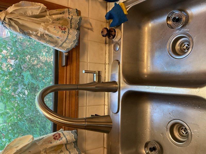 Completed installation of stainless steel double-basin kitchen sink with modern curved faucet assembly featuring dual-handle controls and brushed metallic finish. All fixtures properly mounted in tiled countertop with secure connections and verified drainage functionality in both basins.