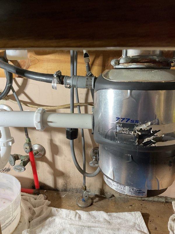 Installed new InSinkErator 777SS garbage disposal unit under kitchen sink, including complete drain assembly with white PVC P-trap configuration, compression fittings, and chrome tailpiece connections. Connected discharge plumbing with proper sealing at all joints, installed water supply shutoff valve with red handle and flexible supply line, and secured all rubber hoses with metal hose clamps to ensure leak-free operation.